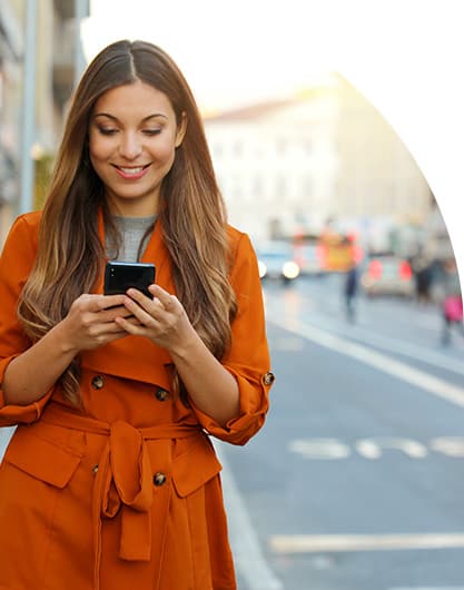 Young adult woman wearing a bright orange jacket and smiling as she completes a service request on her phone while walking down the street.