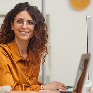 A woman with brown wavy hair wearing glasses and an orange button down shirt working on her computer in a government building.