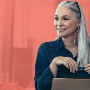 Professional woman in her 60s, against a bright orange background, listening to another person while sitting at her computer.