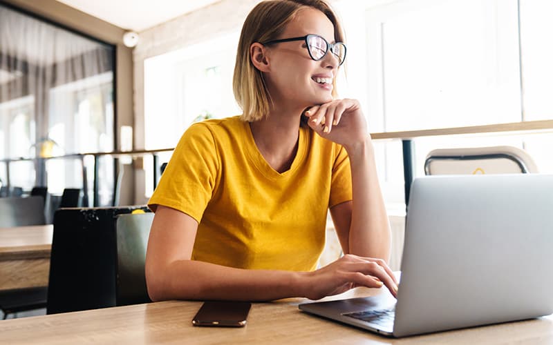 Blonde woman wearing a bright yellow t-shirt smiling while working on laptop.