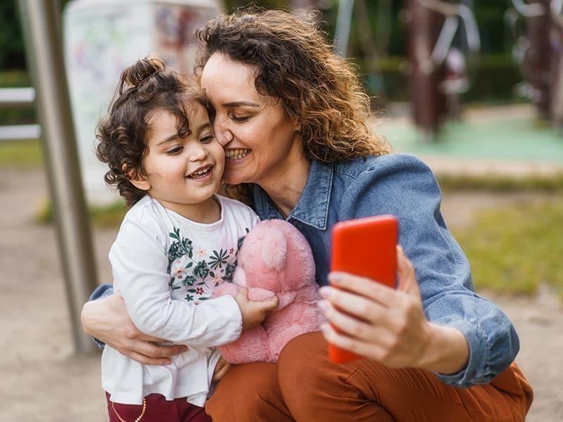 A mother at the park with her young daughter, holding her and her phone while smiling.