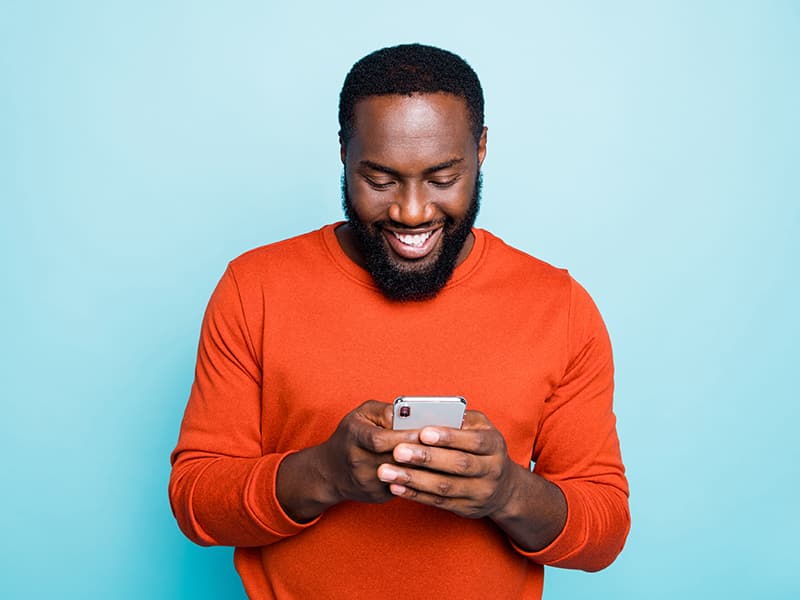 Happy adult man standing against a light blue wall, wearing a bright orange shirt enjoying the simple and easy experience of enrolling in useful government programs on his phone.